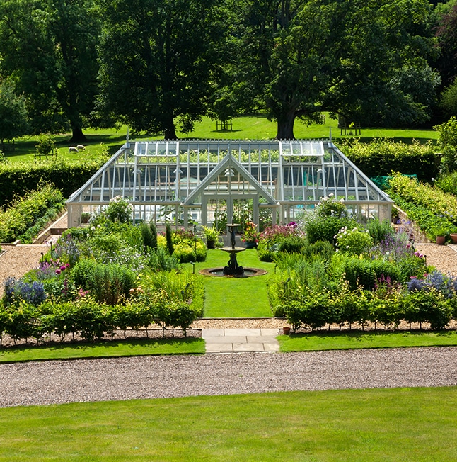 Large Bespoke greenhouse with hipped sides in a large garden containing rows of plants and greenery